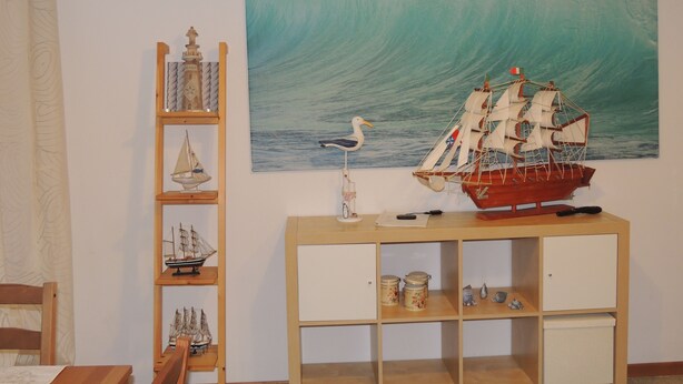 Ein Zimmer mit einem Holztisch, einem Bücherregal mit Modellschiffen, einem großen Gemälde einer Welle und einer Möwenfigur.