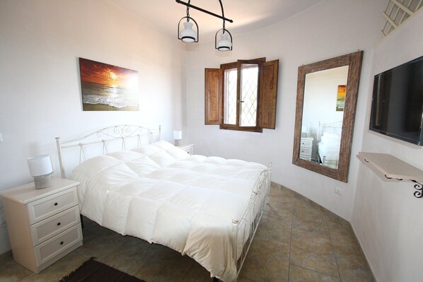 A bedroom with a bed, a nightstand, a lamp, a painting, a window, a mirror, and a TV.