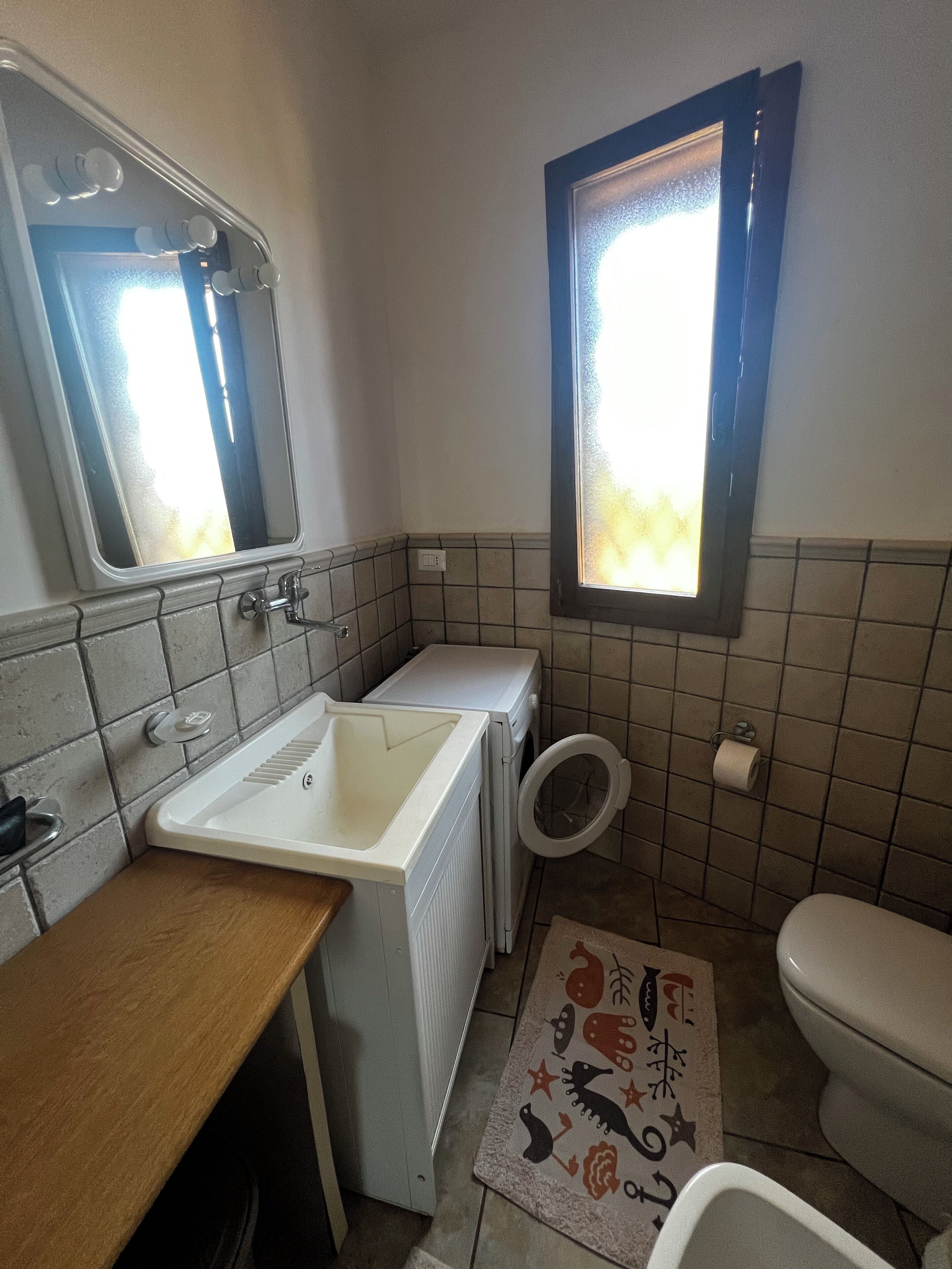 A bathroom with a sink, mirror, toilet, and window.