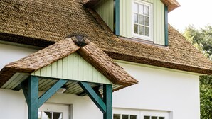 A white building with a green roof, a porch with white chairs, and flower planters. - Small coach house: 10 min. to the beach, in the middle of nature, sauna & whirlpool (Klein Bollhagen)