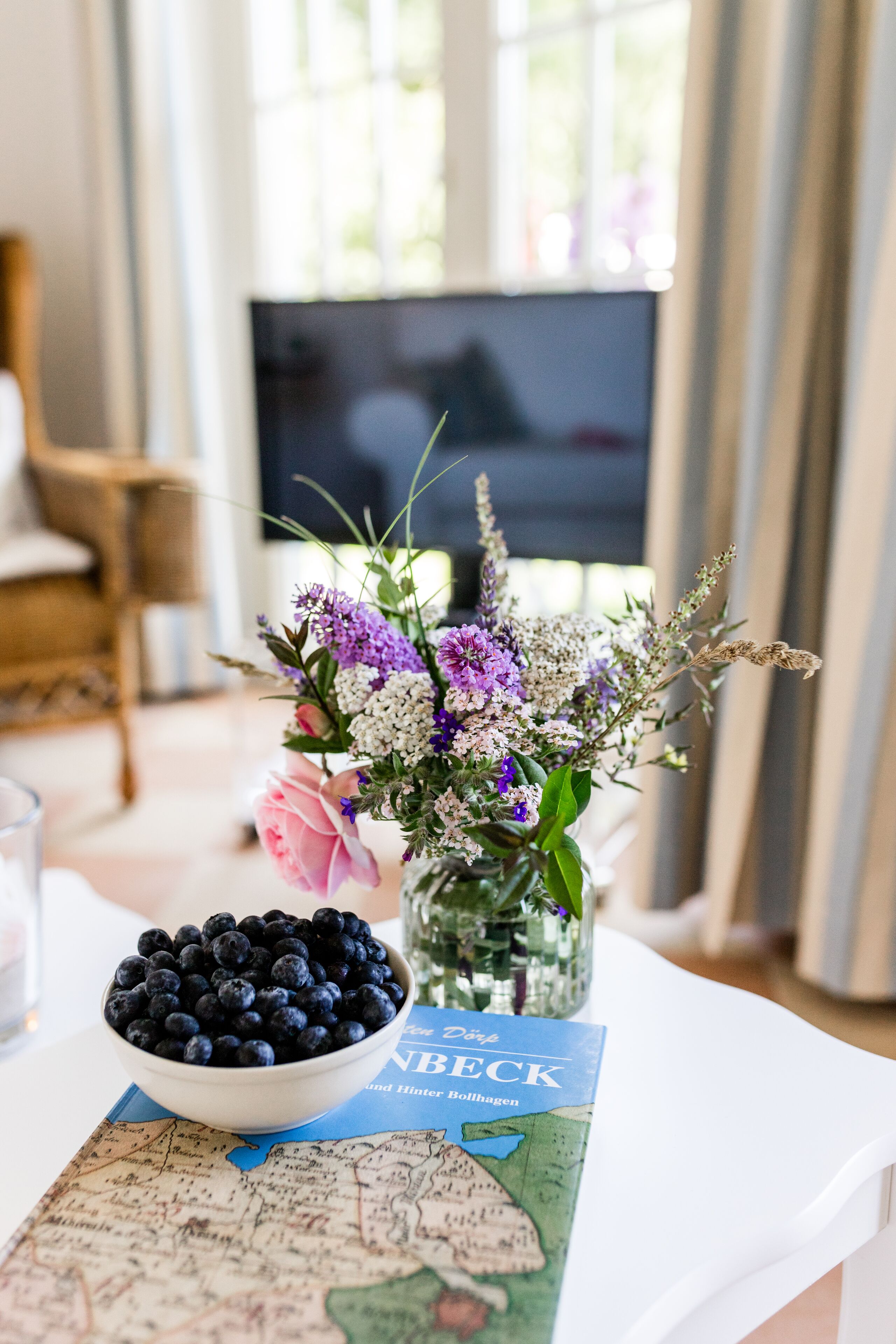 A table with a bowl of blueberries, a vase of flowers, and a map book.