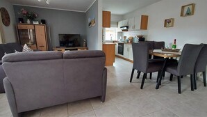 Een woonkamer met een grijze bank, een eettafel met stoelen, een keuken op de achtergrond en een televisie op een dressoir.