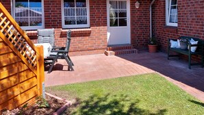 Een bakstenen huis met een rode dakbedekking, een overdekte patio met een houten bankje, een kleine tuin en een houten trap die naar het huis leidt.