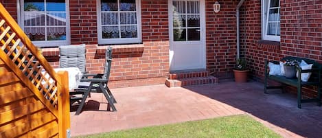 Ein Ziegelhaus mit rotem Ziegeldach, eine überdachte Terrasse mit einer Holzbank, ein kleiner Garten und eine Holztreppe, die zum Haus führt.