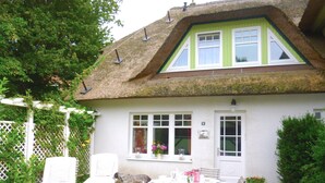 Ein weißes Cottage mit Strohdach, einem Essbereich mit Tisch und Stühlen sowie einem kleinen Garten mit weißem Zaun und viel Grün.