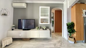 Un salon moderne avec un téléviseur à écran plat posé sur une console blanche, une vitrine en verre contenant des objets décoratifs et une plante en pot.
