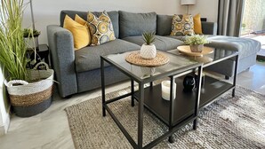Un salon moderne avec un canapé gris, une table basse en verre et des coussins décoratifs. On y trouve également des œuvres d’art murales et une lampe sur pied.