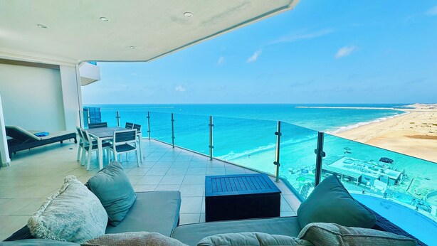 A balcony with a view of the ocean and a beach. There is a table and chairs, and a sofa with cushions.