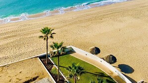 A beach view with a clear sky, calm sea, and a sandy area with palm trees and a lounge area.