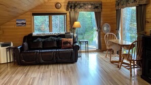L’intérieur de ce chalet chaleureux est doté d’un canapé en cuir, d’une table à manger et de chaises. Une lampe et un ventilateur sont posés sur la table. Les murs de la pièce sont en bois et le plafond est en pente.
