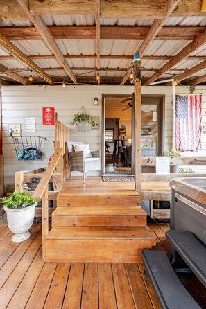 Una terraza de madera con una escalera que lleva a un área cubierta, una zona de estar con una mesa y sillas, y una bandera estadounidense.