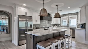 Une cuisine moderne dotée d’un grand îlot central, d’appareils électroménagers en acier inoxydable et de luminaires suspendus.