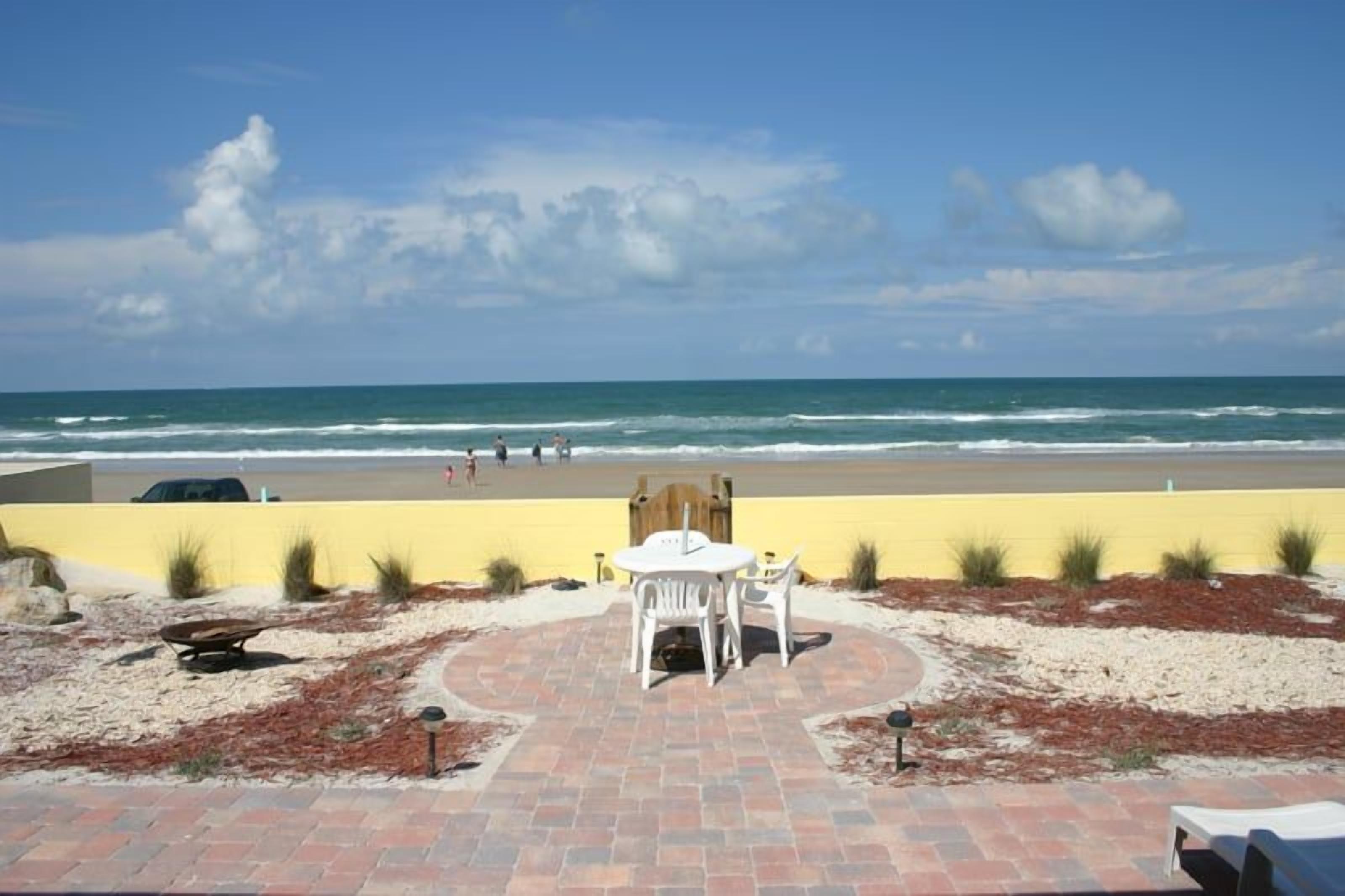 A beachside patio with a table and chairs, a paved walkway, and a view of the ocean.