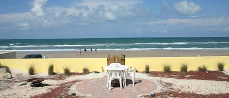 Un patio frente a la playa con mesa y sillas, un sendero pavimentado y vista al océano.