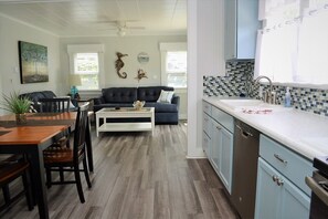 A modern kitchen with a white countertop and a sink, a dining area with a wooden table and chairs, a black leather sofa, and a living room with a dark sofa and a coffee table. - SEAHORSE COTTAGE - Adorable Cottage!! 15 seconds to beach access (Seaside)