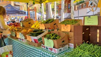 Un puesto de mercado con diversos vegetales y frutas, incluyendo pepinos, calabacines y plátanos, con etiquetas de precio.