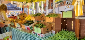 Un puesto de mercado con diversos vegetales y frutas, incluyendo pepinos, calabacines y plátanos, con etiquetas de precio.