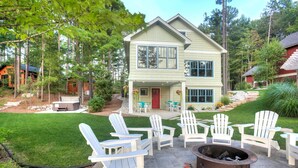 A two-story house with a patio area featuring white Adirondack chairs and a fire pit. - Newly Built Luxury Lakefront Cottage with Fire Pit, Dock, Game Room (Traverse City)