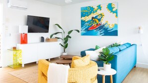 A modern living room with a flat-screen TV, a sofa, a coffee table, and a painting of a surfer. - Spacious Ocean Oasis with Private Outdoor Shower! (Encinitas)