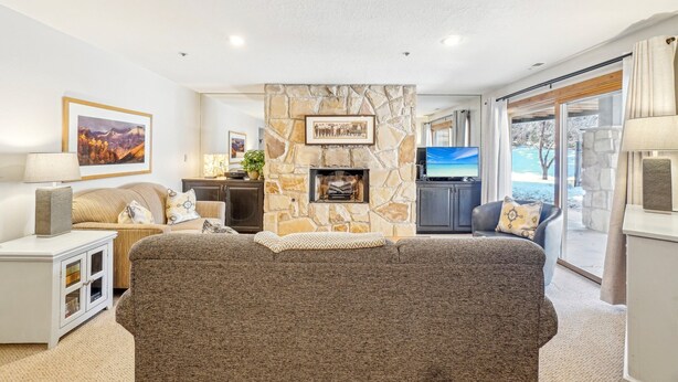 A modern living room with a stone fireplace, a large flat-screen TV, and a view of a pool through the glass door. - All Seasons 2BR | Walk to Park City Mountain | Heated Pool | Sleeps 6 (Park City)
