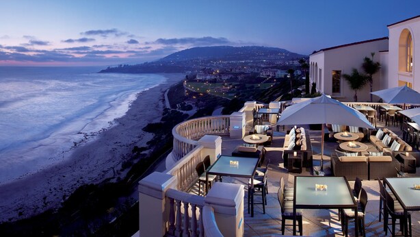 A seaside restaurant with outdoor seating overlooking the ocean, featuring a curved balcony and multiple tables and umbrellas.