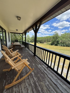 A wooden porch with rocking chairs, a table, and a view of a grassy field and trees.