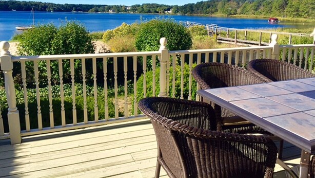 Eine Terrasse mit Korbstühlen und einem Tisch, mit Blick auf einen See mit einer Anlegestelle und Grünflächen.