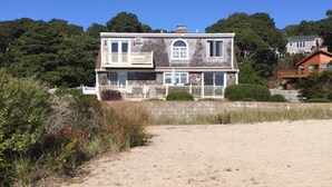 Ein zweistöckiges Haus mit einem sandigen Strand im Vordergrund und dichtem Baumbestand im Hintergrund.