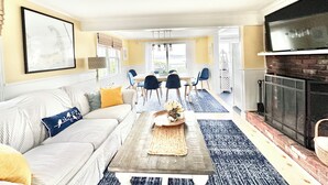 Ein Wohnzimmer mit einer Couch, einem Couchtisch und einem Kamin. Im Hintergrund befindet sich ein Essbereich mit Stühlen und einem Tisch.