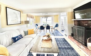 A living room with a sofa, coffee table, and fireplace. There is a dining area with chairs and a table in the background.