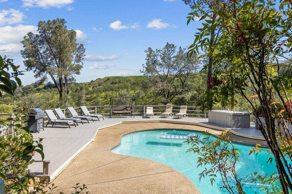 A pool area with lounge chairs and a hot tub, surrounded by trees and hills.