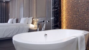 A modern bathroom with a freestanding bathtub, a large mirror, and a bed with white linens in the background. - The Mira Central Park Hotel (Bien Hoa)