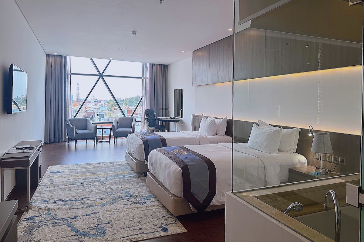 A modern hotel room with a large bed, a desk, a chair, and a view of the city through large windows.