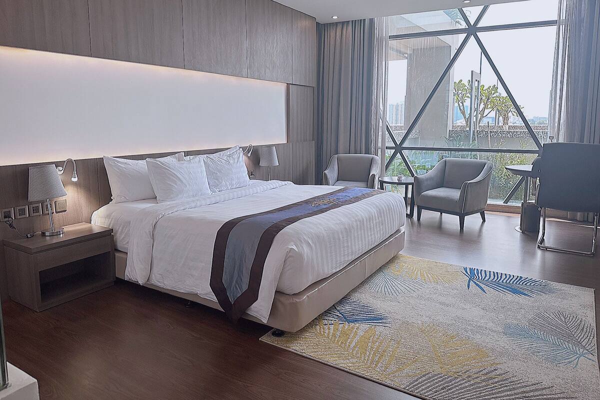 A modern hotel room with a large bed, two armchairs, a small table, and a view of the city through large windows.