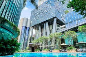 A modern building with a geometric facade, a pool area with lounge chairs and umbrellas, and lush greenery. - The Mira Central Park Hotel (Bien Hoa)