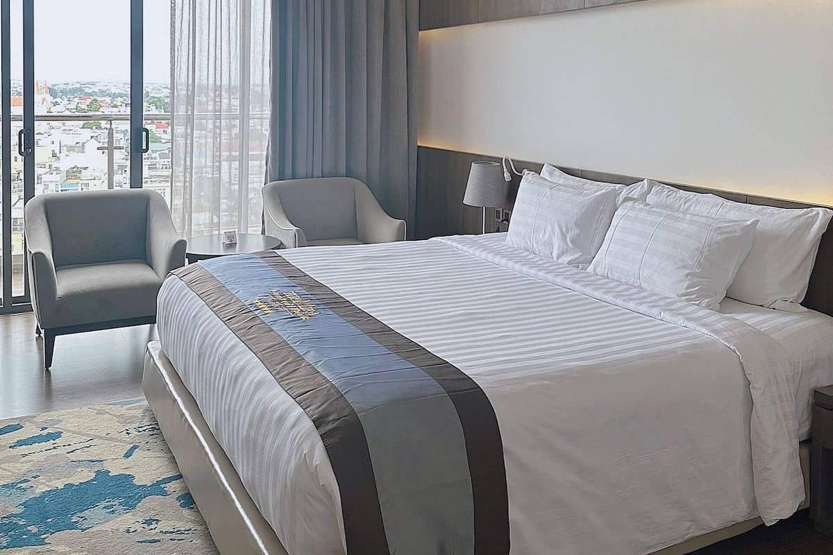 A modern hotel room with a large bed, two armchairs, a view of the city through large windows, and a balcony.