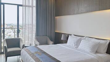 A modern hotel room with a large bed, two armchairs, a view of the city through large windows, and a balcony.