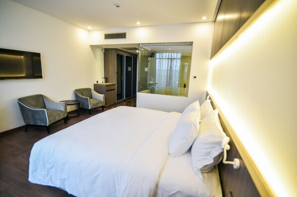 A hotel room with a large bed, two armchairs, a small table, a mirror, and a shower area.