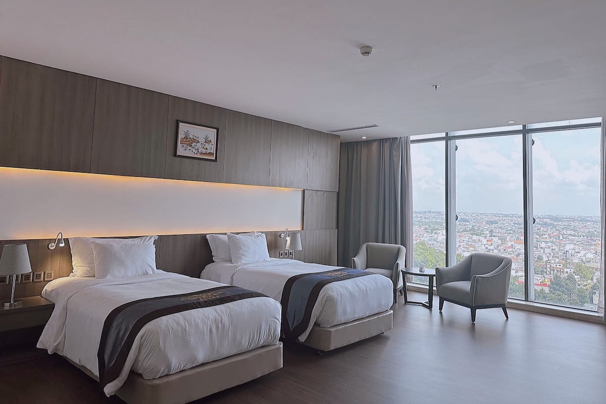 A hotel room with two beds, a chair, a small table, and a large window with a city view.