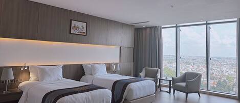A hotel room with two beds, a chair, a small table, and a large window with a city view.