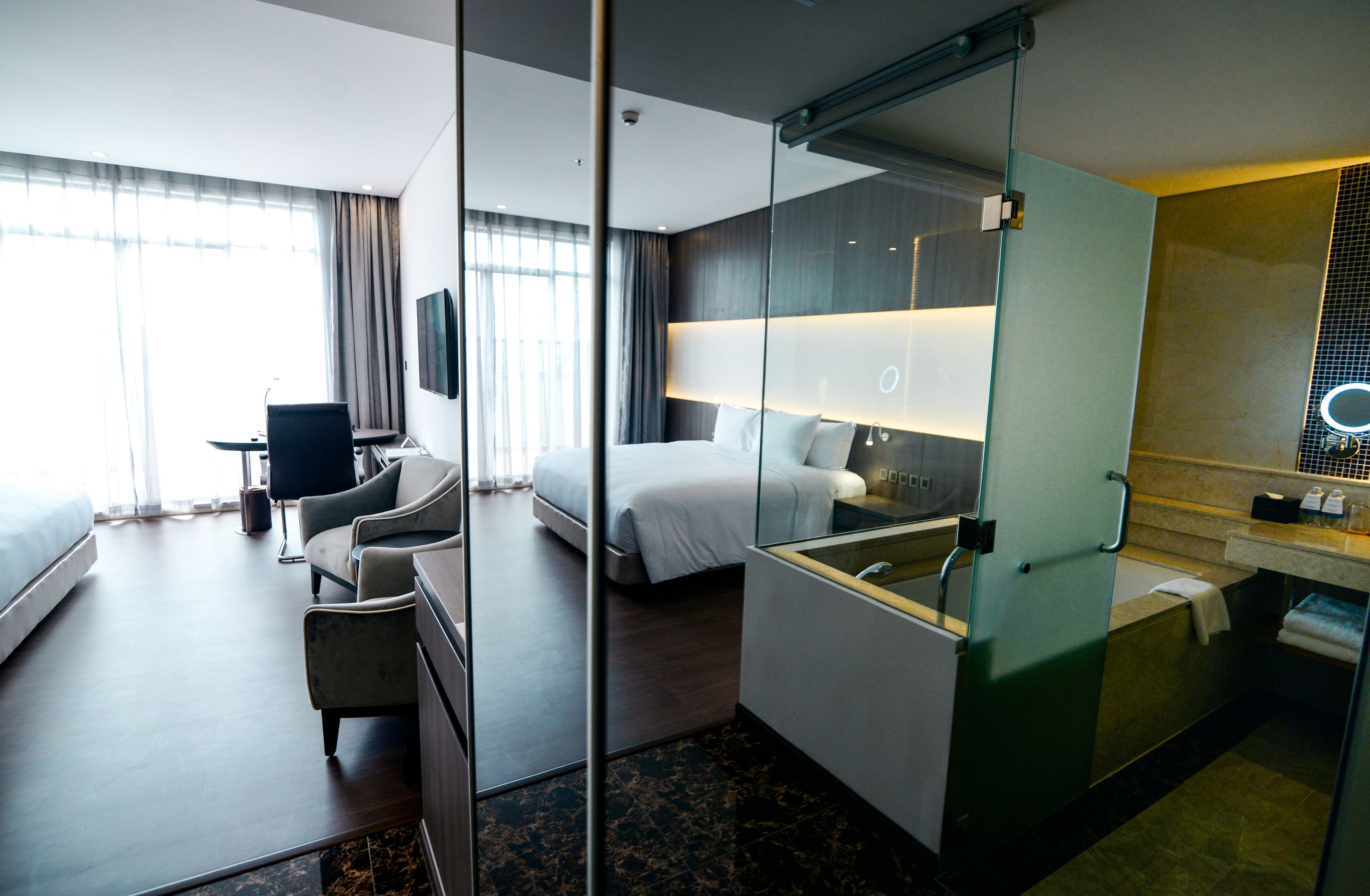 A modern hotel room with a bed, a desk, a chair, a glass partition, and a bathroom with a mirror and sink.