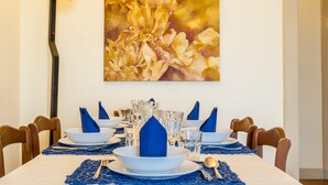 Ein Esstisch ist gedeckt mit blauen Servietten, weißen Tellern und klaren Gläsern, über der Tischwand hängt ein großes Blumenbild.