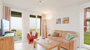 Un salon spacieux avec un grand canapé, une table basse en bois et un téléviseur posé sur un meuble en bois. On y trouve également deux fauteuils et une grande fenêtre donnant sur l’extérieur.