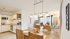 Une cuisine moderne avec des meubles blancs, un four à micro-ondes et une machine à café. On y trouve un coin repas avec une table et des chaises en bois, et une vue sur la mer grâce à de grandes fenêtres.