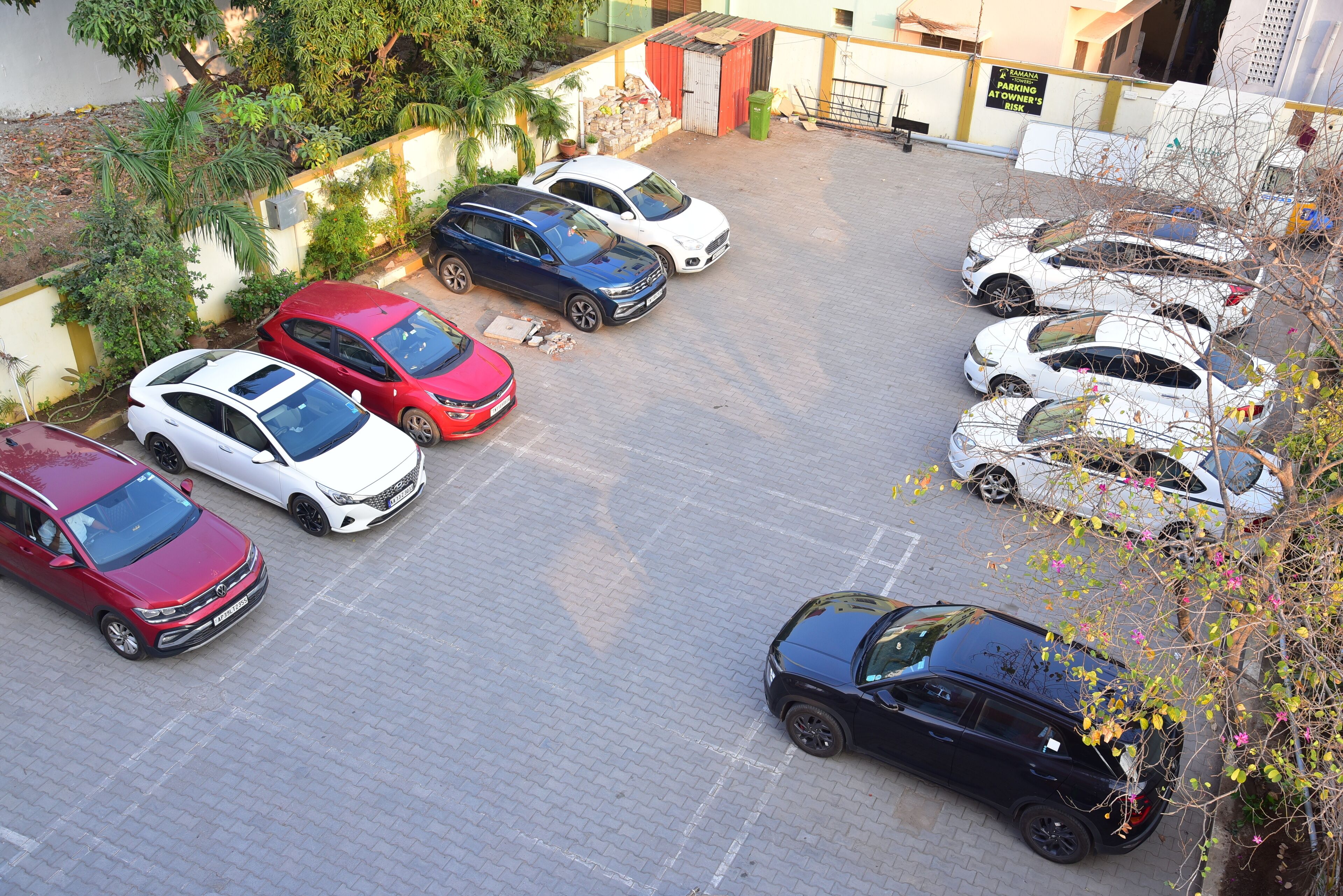 Un estacionamiento con varios autos, un edificio de fachada amarilla y blanca, y algo de vegetación.