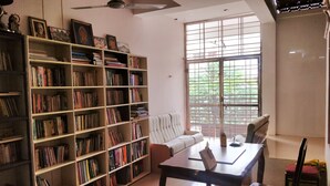 A room with a large bookshelf filled with books, a sofa, a table with chairs, and a window with blinds. - Ramana Towers (Tiruvannamalai)