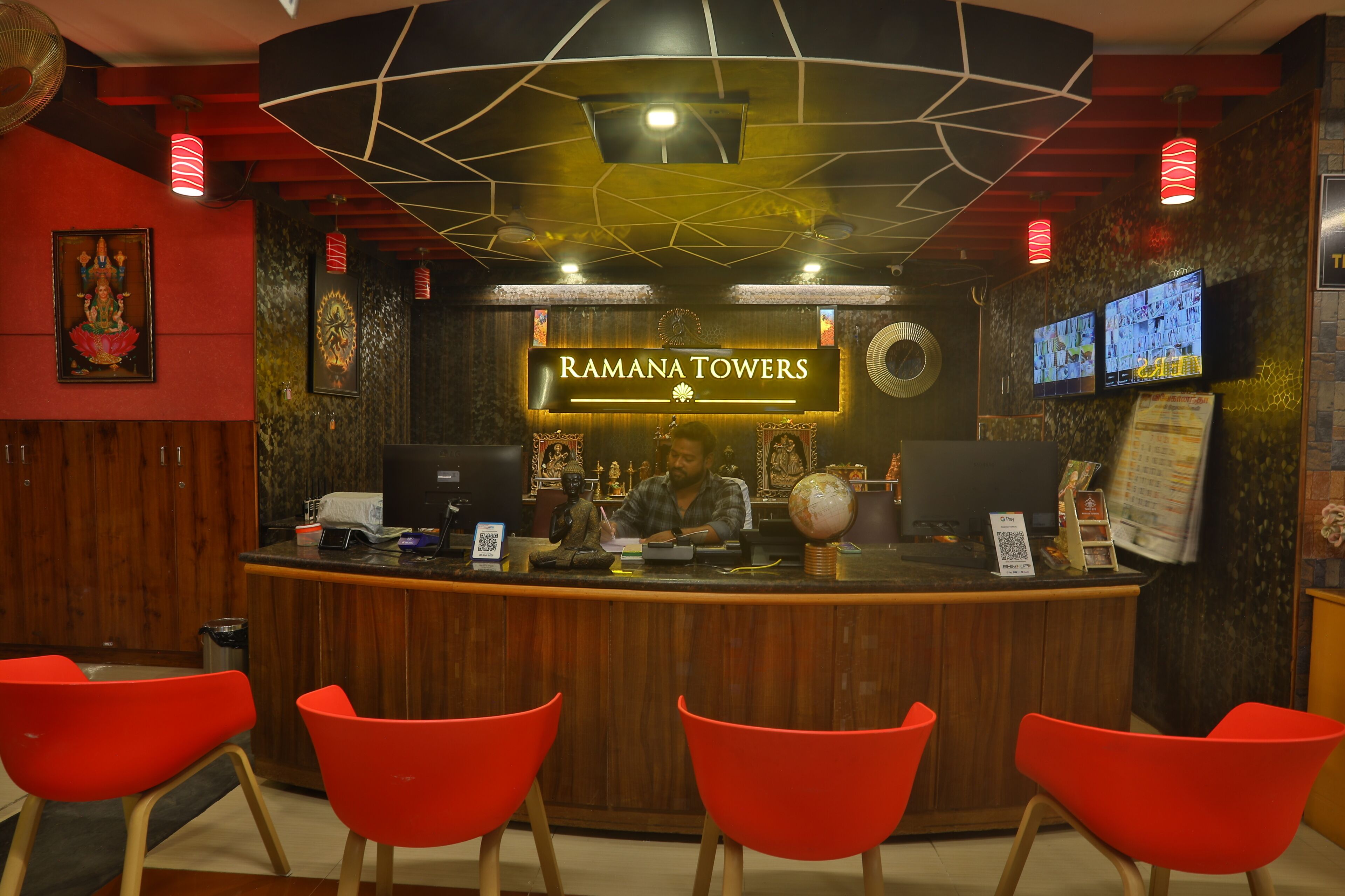 A reception desk with a sign reading 'RAMANA TOWERS' and a Buddha statue. There are red chairs in front of the desk.