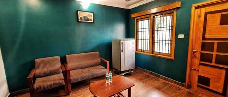 Una habitación con paredes verde azulado, una puerta de madera, una ventana con barrotes, dos sillas estampadas, una mesita y un refrigerador.