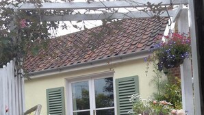 Une maison avec un toit en tuiles, une porte blanche et des volets verts. Des marches mènent à la porte, et des plantes en pot, ornées de fleurs, sont disposées à l’extérieur.