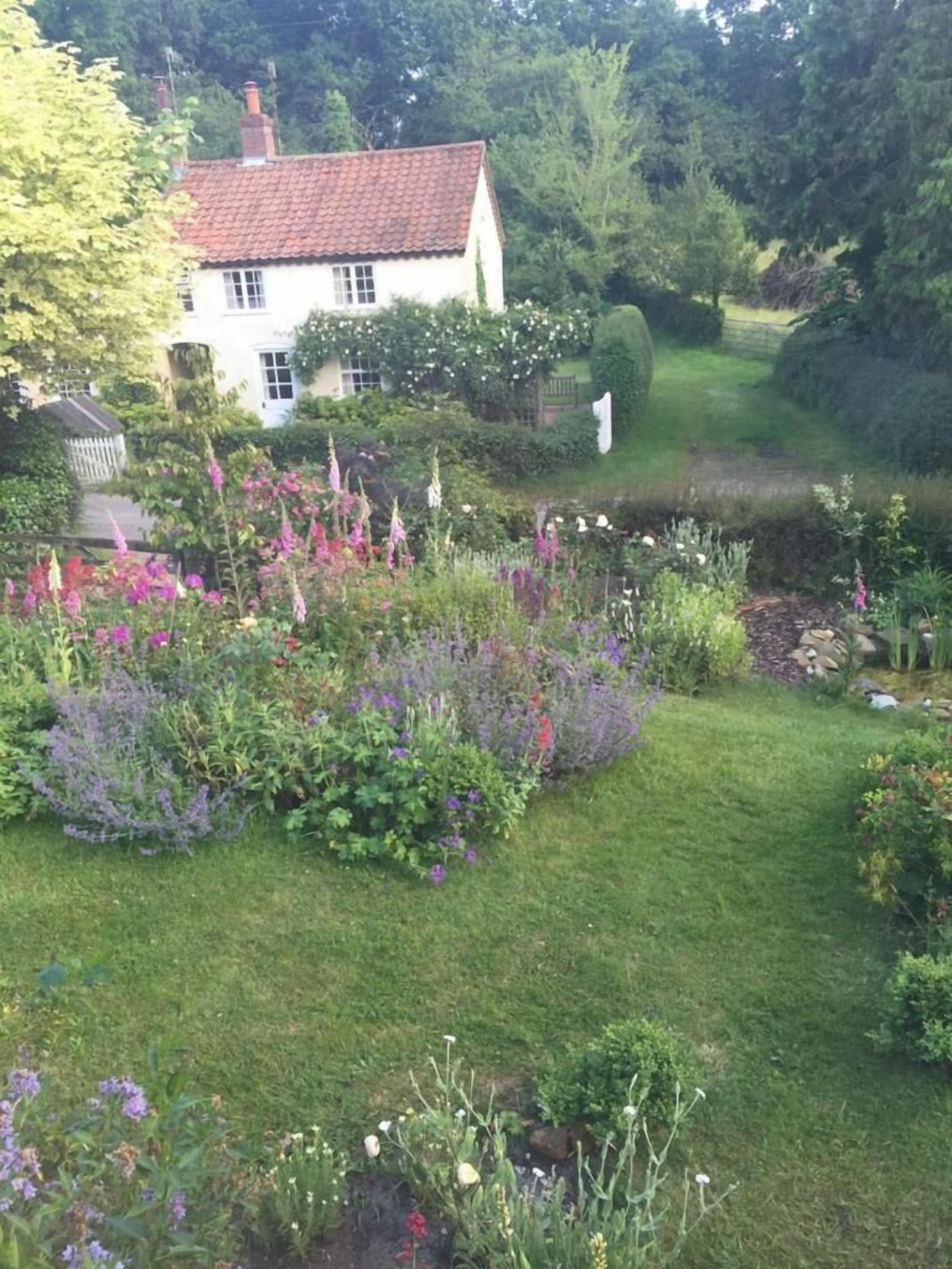 A garden with a variety of plants and flowers, a white house with a red roof, and a pathway leading to the house.